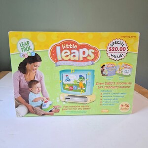 Puede incluir: Una caja del sistema de aprendizaje LeapFrog Little Leaps Grow-with-Me. La caja es verde, amarilla y blanca, con imágenes de una madre y un bebé, y un juguete con forma de televisor. Texto: "Share baby's discoveries!" y "9-36 meses."