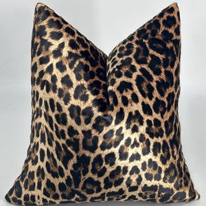 May include: A square cushion with a leopard print design. The cushion has a tan base with dark brown spots, creating a classic animal print. The fabric appears soft. The cushion would suit a living room or bedroom.