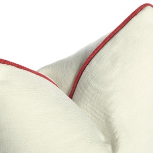 May include: Two white throw pillows with red piping around the edges. The pillows are made of a textured fabric and are arranged in a way that shows the piping.