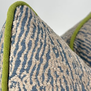 May include: A decorative throw pillow featuring a textured fabric with a blue and beige abstract pattern. The pillow is finished with a bright lime green piping along the edge, providing a distinct contrast to the blue and beige woven material.