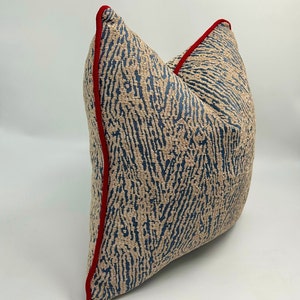 Op de afbeelding: Een blauw en beige kussen met een patroon en een rode rand. Het kussen heeft een textuur en een geometrisch ontwerp.