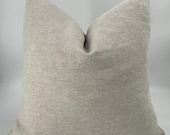 May include: A square throw pillow featuring a textured, light beige fabric with a subtle woven pattern. The pillow has a plump, filled appearance with defined corners and a consistent, neutral-toned surface throughout the entire visible exterior.