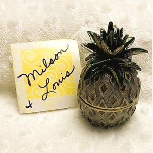 May include: A gold pineapple-shaped trinket box with a green top and a diamond pattern on the body. The box is sitting on a white surface with a yellow card that says "Milson & Louis" on it.
