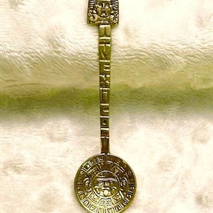 May include: A gold-colored souvenir spoon with an Aztec calendar design on the bowl. The handle features a carved face and the word "VENEZ" is etched on the handle. The spoon is approximately 6 inches long.