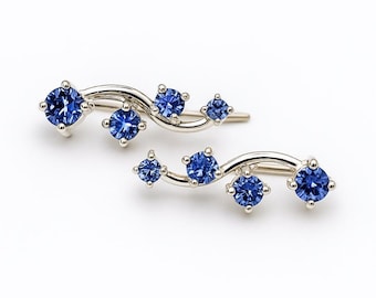 Puede incluir: Un par de trepadores de oreja plateados, cada uno con un diseño curvo adornado con cinco piedras preciosas azules brillantes. Las piedras preciosas varían en tamaño, creando una estética fluida y elegante. Los pendientes están sobre un fondo blanco.