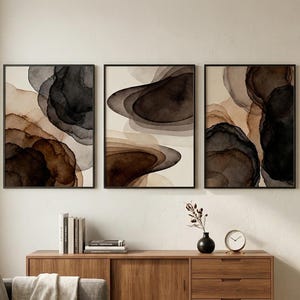 Peut inclure: Trois impressions d'art abstraites encadrées. Chaque impression présente des formes de style aquarelle dans des tons de marron, noir et beige. L'œuvre est exposée au-dessus d'un meuble en bois. Une plante et une horloge sont également visibles.