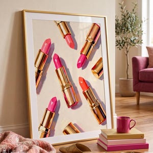 May include: A framed wall art print featuring a pattern of multiple lipsticks in gold tubes with various shades of pink and orange lipstick bullets, displayed against a light cream background within a thin gold frame.