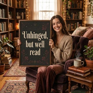 May include: A framed sign with the text "Unhinged, but well read" held by a person. The sign has a dark background with floral accents. The person is seated in a velvet chair in a room filled with books.