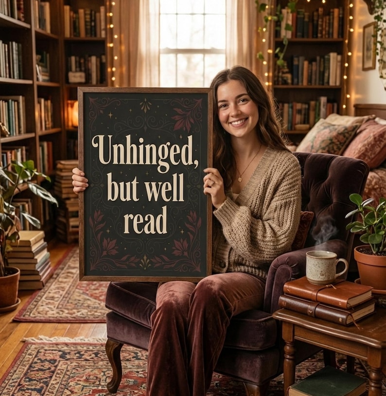 May include: A framed sign with the text "Unhinged, but well read" held by a person. The sign has a dark background with floral accents. The person is seated in a velvet chair in a room filled with books.