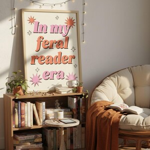 May include: A framed print with the text "In my feral reader era" in pink and orange lettering. The print is displayed above a wooden bookshelf filled with books. A cozy reading nook is set up with a chair and blanket.