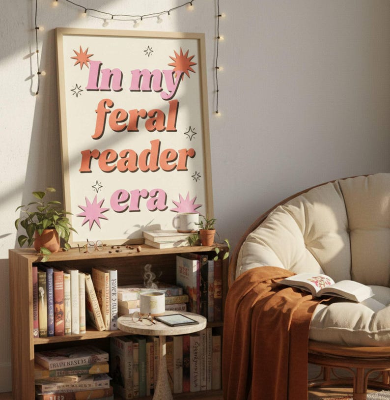 May include: A framed print with the text "In my feral reader era" in pink and orange lettering. The print is displayed above a wooden bookshelf filled with books. A cozy reading nook is set up with a chair and blanket.