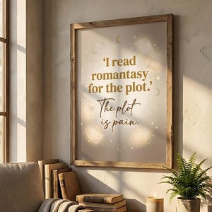 May include: Wooden framed print with the quote, "I read romantasy for the plot. The plot is pain." The print has a cream background with illustrations of books and stars. The print is displayed on a wall next to a window and a stack of books.
