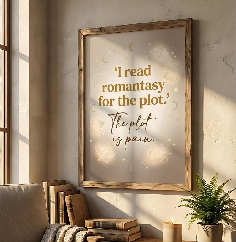May include: Wooden framed print with the quote, "I read romantasy for the plot. The plot is pain." The print has a cream background with illustrations of books and stars. The print is displayed on a wall next to a window and a stack of books.