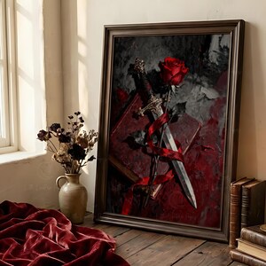 Puede incluir: Obra de arte enmarcada que presenta una rosa roja, una espada y un libro, todos entrelazados con una cinta roja. El fondo es una mezcla de tonos rojo oscuro y gris. La obra est&aacute; en un marco marr&oacute;n oscuro, colocada sobre una superficie de madera.