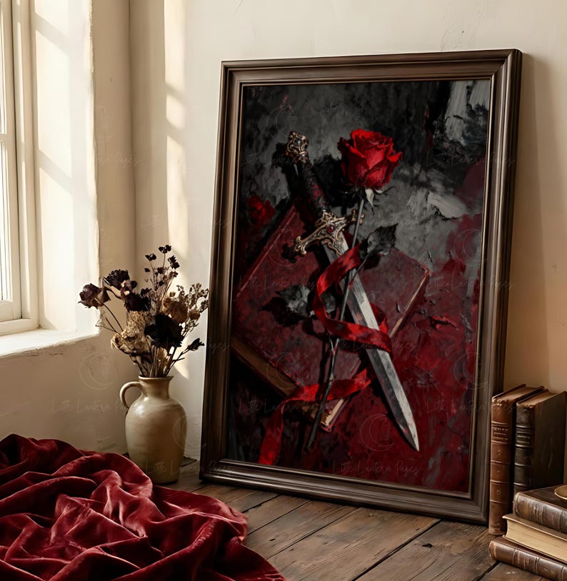 Puede incluir: Obra de arte enmarcada que presenta una rosa roja, una espada y un libro, todos entrelazados con una cinta roja. El fondo es una mezcla de tonos rojo oscuro y gris. La obra est&aacute; en un marco marr&oacute;n oscuro, colocada sobre una superficie de madera.