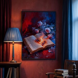 May include: A framed artwork depicts an open book with flames at its center, surrounded by roses and ice crystals against a red and blue background. A blue lamp with a pleated shade sits on a wooden table next to the artwork.