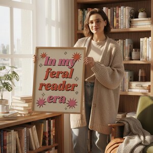 May include: A framed poster with the text "In my feral reader era" in pink and orange lettering. The poster is held by a person in a beige cardigan, standing in front of a bookshelf filled with books. The poster has star and sunburst designs.