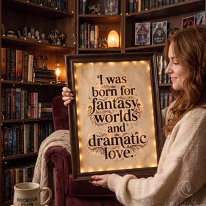 May include: A rectangular dark wood frame containing a parchment-colored sign with the black text: I was born for fantasy worlds and dramatic love. The sign is illuminated by a string of small warm white LED lights lining the inner frame edge.