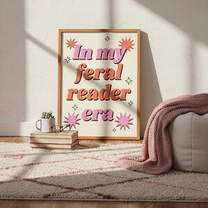 May include: A framed print with the text "In my feral reader era" in pink and orange retro font. The print is in a light brown frame and is decorated with starburst and star graphics. The print is on a cream background.