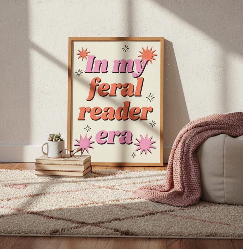 May include: A framed print with the text "In my feral reader era" in pink and orange retro font. The print is in a light brown frame and is decorated with starburst and star graphics. The print is on a cream background.