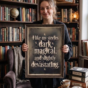 Op de afbeelding: Een ingelijste print met de tekst "I like my stories dark, magical, and slightly devastating." De print heeft een donkere achtergrond met witte letters en decoratieve elementen. De lijst is bruin. De afbeelding wordt voor een boekenplank gehouden.