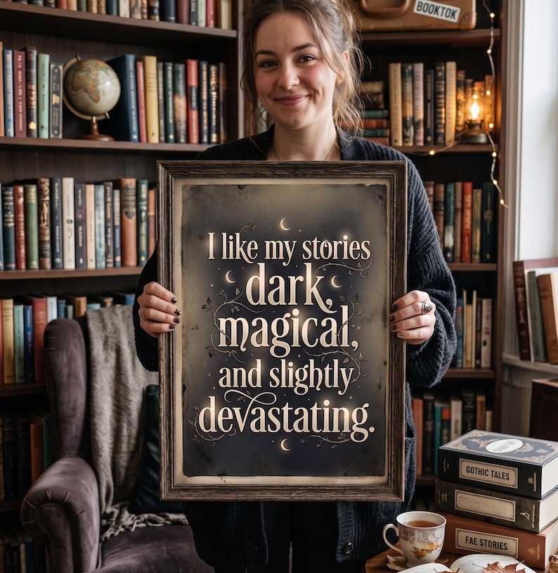 Op de afbeelding: Een ingelijste print met de tekst "I like my stories dark, magical, and slightly devastating." De print heeft een donkere achtergrond met witte letters en decoratieve elementen. De lijst is bruin. De afbeelding wordt voor een boekenplank gehouden.