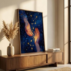 May include: A framed art print featuring a celestial theme. The artwork depicts two hands reaching towards each other against a dark blue background with golden stars and constellations. The hands are illuminated with a warm, golden light.