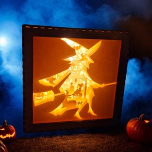 May include: A wooden shadow box with a cut-out design of a witch flying on a broomstick. The box is lit from behind, creating a glowing effect. The witch is holding a cauldron and there are other Halloween-themed elements in the design.