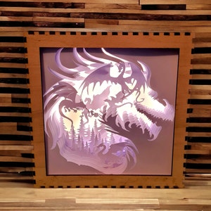 Pode incluir: Uma moldura de madeira com uma silhueta de dragão roxo claro dentro. O dragão é feito de papel em camadas e é iluminado por trás. O dragão está em um cenário de floresta.