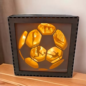 Puede incluir: Un marco de madera negro con un diseño de balón de fútbol 3D iluminado. El balón está hecho de papel en capas y presenta siluetas de jugadores de fútbol. El marco está iluminado con luces LED blancas cálidas.