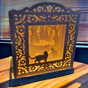 May include: A black papercraft lamp with a silhouette of cats in a forest scene. The lamp is illuminated from within, casting a warm glow.