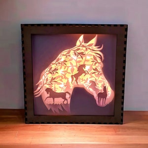 Puede incluir: Una obra de arte en capas con la forma de una cabeza de caballo en un marco marrón oscuro. La cabeza del caballo presenta siluetas de hojas y caballos, iluminadas con luz cálida. Una pieza de decoración del hogar única.