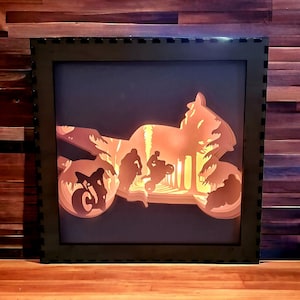 May include: A black framed wall art piece with a silhouette of a motorcycle and riders. The motorcycle is made of wood and is lit from behind, creating a warm glow.