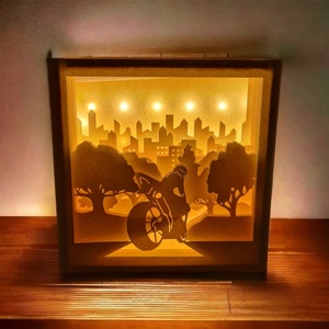 Può includere: Una scatola luminosa in carta 3D con una silhouette di città e un motociclista. La scatola è illuminata dall'interno, creando un bagliore caldo.