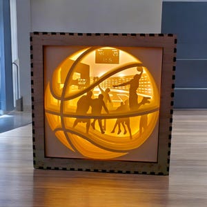 Op de afbeelding: Een gelaagd papieren kunstwerk dat een basketbalspelscène uitbeeldt. Het kunstwerk is ingelijst in een houten frame en heeft een basketbalontwerp. Het verlichte interieur toont spelers, een scorebord en een basket, waardoor een warme, uitnodigende gloed ontstaat.