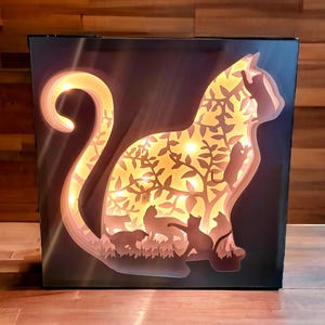 Könnte beinhalten: Dekorative katzenförmige Leuchtbox mit mehrschichtigem Design. Die Silhouette einer Katze ist mit einer Waldszene gefüllt, einschließlich Bäumen und anderen Katzen. Die Leuchtbox strahlt ein warmes Licht aus und erzeugt eine gemütliche Atmosphäre. Die Box ist schwarz mit einem Holzsockel.