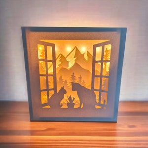Könnte beinhalten: Eine Holz-Schattenbox mit einer Fensterszene, die zwei Katzen zeigt, die sich gegenseitig ansehen. Die Szene ist mit warmweißen LED-Lichtern beleuchtet und enthält eine Bergkette und Bäume.
