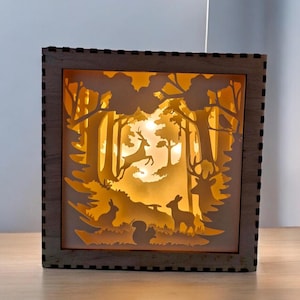 Peut inclure: Une boîte lumineuse carrée en bois représentant une forêt en couches. La conception comprend des silhouettes d'arbres, de cerfs, d'un lapin et d'un écureuil, éclairées par une lumière jaune chaude. Le cadre a un bord sombre et pointillé.