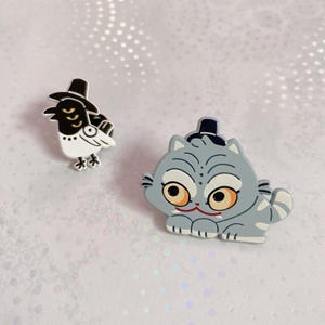 May include: Two enamel pins featuring cartoon animals. One pin is a black and white bird wearing a black top hat. The other is a gray cat with orange eyes and a black top hat. Both pins have a whimsical design.