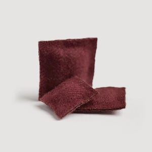May include: Three burgundy-colored, square-shaped felt pieces. One is upright, and two are lying flat, creating a layered effect. The felt has a textured surface, and the color is a deep, rich shade of red.