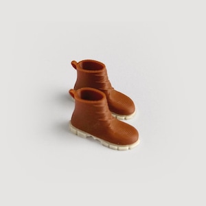 May include: A pair of miniature brown boots with white soles. The boots have a textured surface and a small loop at the top. The boots are a warm brown color and are set against a white background.