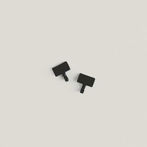 May include: Two small, black, T-shaped objects are positioned on a neutral background. The objects have a rectangular top and a cylindrical stem. The items appear to be keys or similar small hardware components.