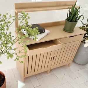 Könnte beinhalten: Ein hellbraunes Holzkabinett mit zwei Türen und einer Schublade. Das Kabinett hat ein Regal oben und ist mit vertikalen Latten verziert. Es befinden sich drei Topfpflanzen auf dem Regal und dem Boden.