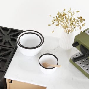 Puede incluir: Una escena de cocina en miniatura con cuencos blancos con bordes negros, una pequeña cuchara de madera y un jarrón decorativo con flores secas. También se ven una máquina de espresso verde y una estufa negra. La escena está ambientada en una encimera blanca.