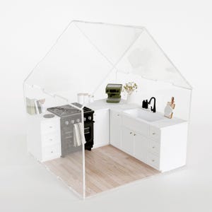 Peut inclure: Maquette de cuisine miniature avec des meubles blancs, une cuisinière noire et un évier. Le modèle comprend une petite cafetière, une planche à découper et un vase de fleurs. La cuisine est enfermée dans une structure transparente en forme de maison.