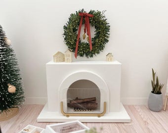 May include: White miniature fireplace with a glass fire screen and a wreath with red ribbon and gold pine cones hanging above. The fireplace is decorated with small wooden houses.
