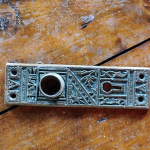 May include: A brass door knob with an ornate design and a keyhole. The knob is rectangular and has a decorative border.
