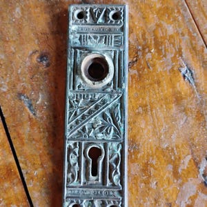 May include: Antique brass door plate with intricate floral and geometric designs. The plate has a keyhole and a round hole for a knob.