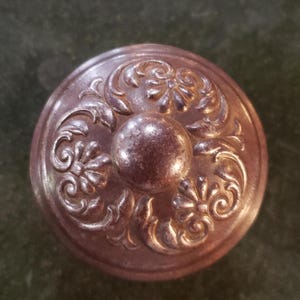 May include: A round, ornate, metal knob with a raised, circular center and intricate floral designs. The knob is a dark brown color with a metallic sheen.