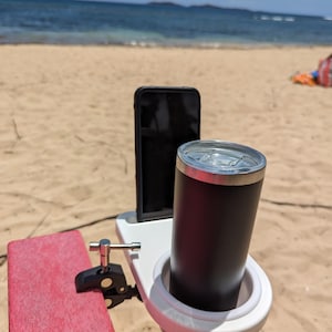 May include: A white phone and drink holder with a black clamp attached to a red surface. The holder is holding a black phone and a black tumbler. The holder is on a beach with a blue ocean in the background.